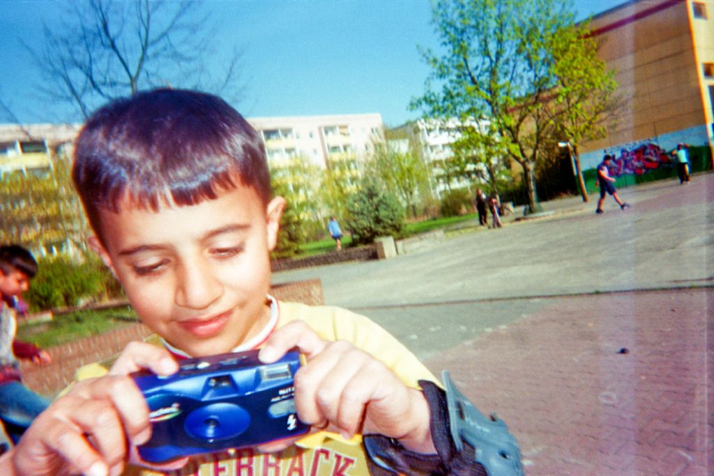 Das farbenfrohe Bild eines kleinen Jungen, der lächelnd auf seine blaue Einwegkamera schaut.