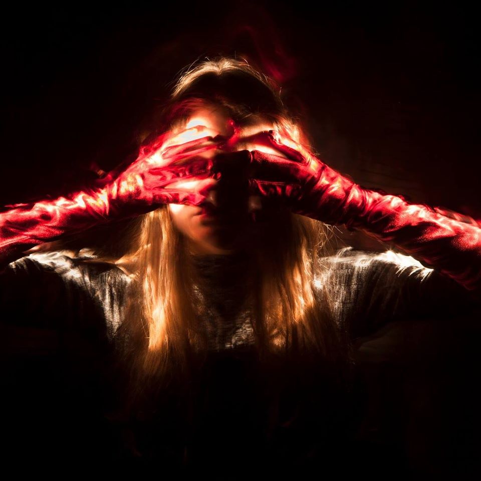 Farbfotoportrait einer jungen Frau mit langem blonden Haar, die sich beide Hände quer vor die Augen hält. Die Arme stecken in langen roten Handschuhen, durch die Lightpaintingtechnik scheint das Licht zwischen ihren Fingern zu scheinen.